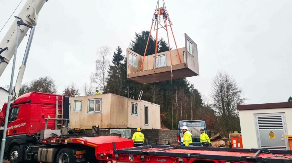 Ein Kran hebt einen modularen Wohncontainer mit Fenstern auf einer Baustelle.